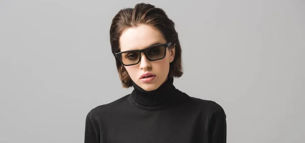 Colpo panoramico di attraente giovane donna in maglione nero e occhiali da sole isolati su grigio — Foto stock