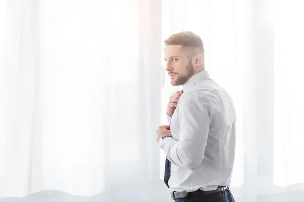 Красивий бородатий чоловік торкається краватки, стоячи вдома — стокове фото