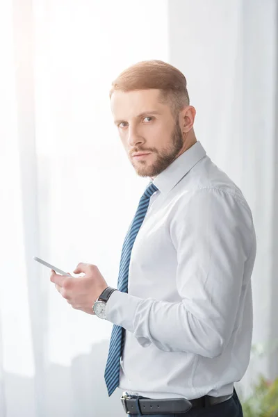 Handsome bearded man in suit holding smartphone while looking at camera — Stock Photo