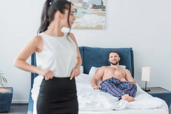 Enfoque selectivo del hombre barbudo acostado en la cama y mirando a la mujer morena en el dormitorio - foto de stock