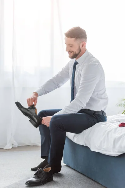 Hombre barbudo alegre en ropa formal sentado en la cama y sosteniendo el zapato - foto de stock