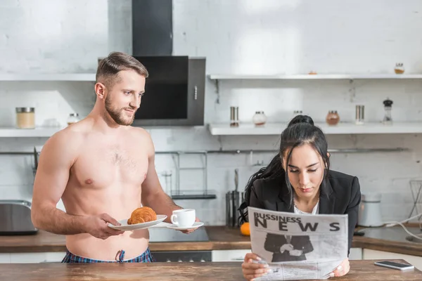 Bell'uomo senza camicia che tiene il piattino con croissant e tazza di caffè vicino alla donna bruna che legge il giornale aziendale — Foto stock