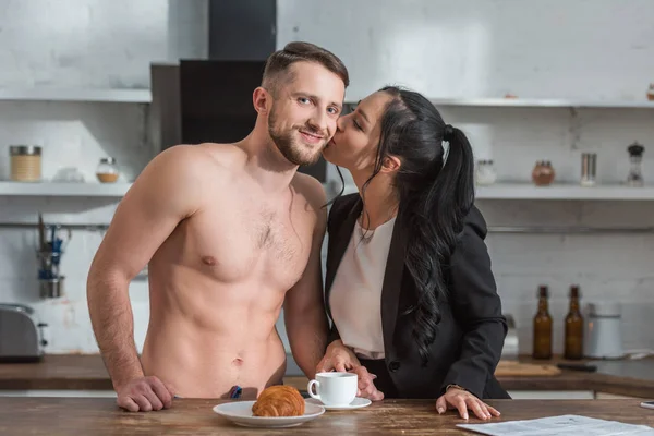 Morena mujer besando mejilla de feliz hombre sin camisa cerca de taza de café y croissant - foto de stock