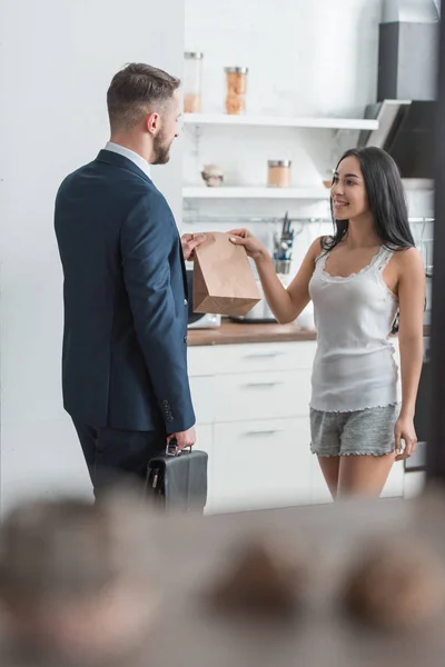 Selective focus of cheerful brunette woman giving paper bag to handsome man in suit — Stock Photo