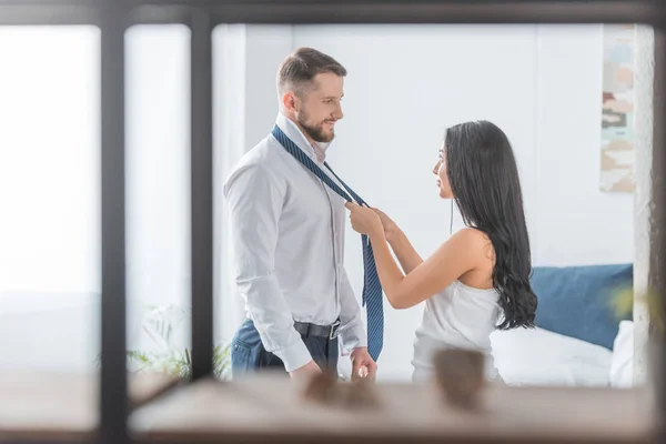 Selective focus of beautiful brunette woman tying tie on bearded man at home — Stock Photo
