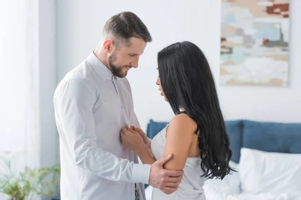 Brünette junge Frau berührt im Schlafzimmer stehenden hübschen Freund mit weißem Hemd — Stockfoto