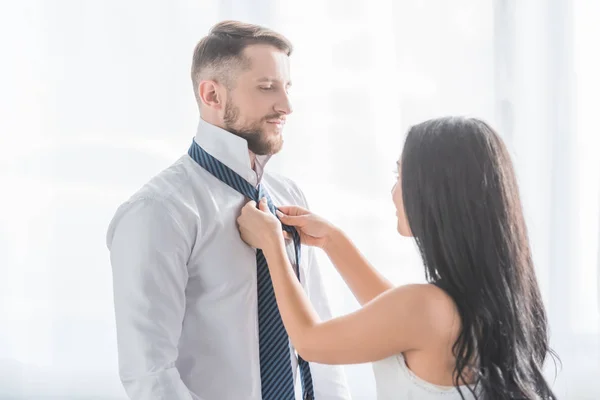 Брюнетка дівчина зв'язує краватку красивого бородатого чоловіка в білій сорочці — стокове фото