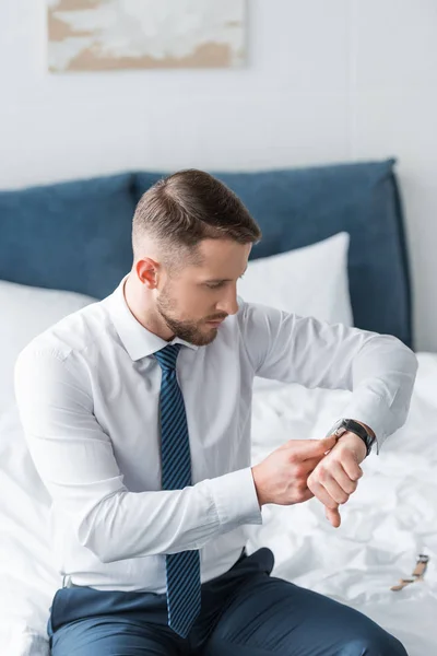 Bello uomo barbuto in abbigliamento formale guardando orologio mentre seduto sul letto — Foto stock