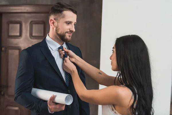 Atractiva chica morena tocando corbata de hombre guapo de pie en traje con rollo de papel - foto de stock