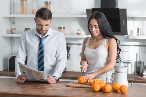Atractiva chica morena cortando naranja en tabla de cortar cerca de hombre leyendo periódico - foto de stock