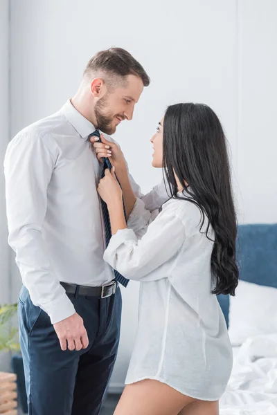 Feliz novia en camisa blanca tocando corbata de guapo novio en traje - foto de stock