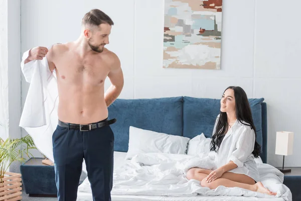 Attractive girl sitting on bed and looking at handsome man wearing white shirt — Stock Photo