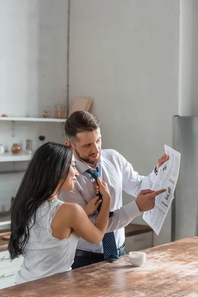 Cheerful woman touching tie of handsome man in suit reading newspaper in kitchen — Stock Photo