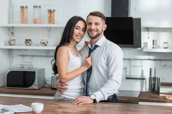 Alegre chica tocando corbata de guapo novio en traje mientras de pie en cocina - foto de stock