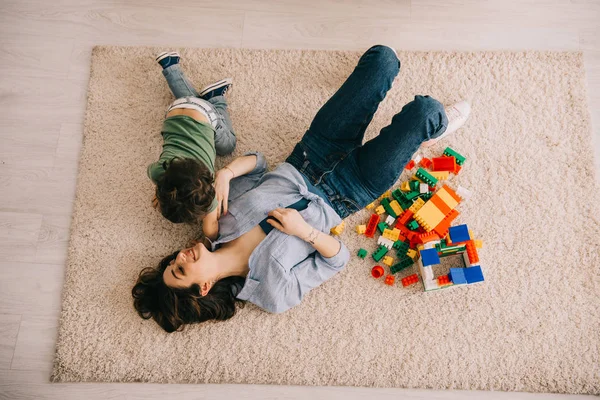 Над головой вид улыбающихся мамы и сына, лежащих на ковре с игрушечными блоками — стоковое фото