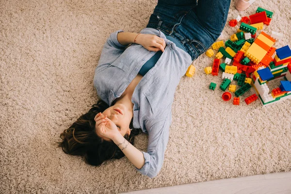 Visão overheaf de mulher cansada deitada no tapete com blocos de brinquedos coloridos — Fotografia de Stock