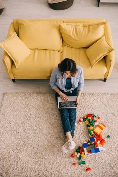 Vista aérea da mulher sentada no tapete perto de blocos de brinquedos e usando laptop — Fotografia de Stock