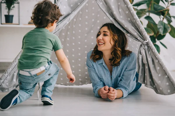 Sonriente madre e hijo en wigwam gris en casa - foto de stock