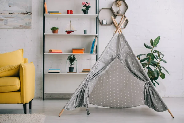 Wigwam gris cerca de estantes con libros y sofá amarillo - foto de stock