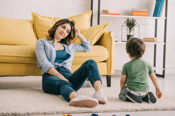 Müde Mutter sitzt auf Teppich und schaut Sohn an — Stockfoto