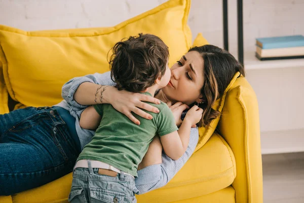 Маленький мальчик целует уставшую мать, лежащую на желтом диване — стоковое фото
