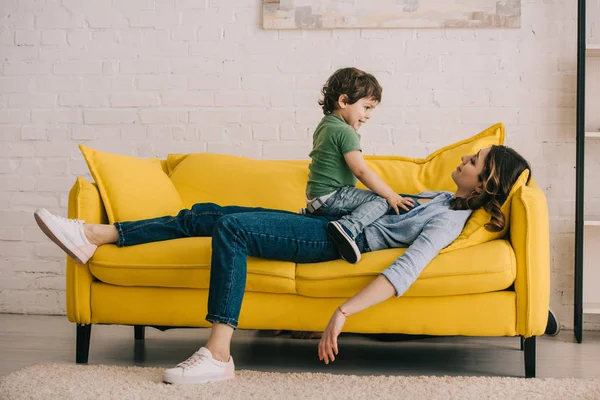 Pequeño niño sentado en la madre cansada mientras ella acostada en el sofá amarillo en la sala de estar - foto de stock