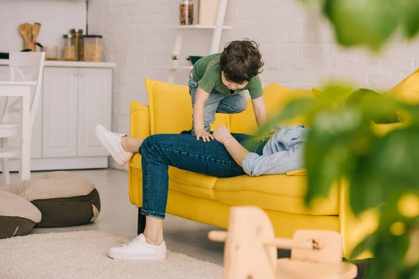 Little boy playing with tired mother while she lying on yellow sofa in living room — Stock Photo