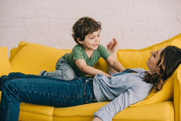 Little boy sitting on tired mother while she lying on yellow sofa in living room — Stock Photo