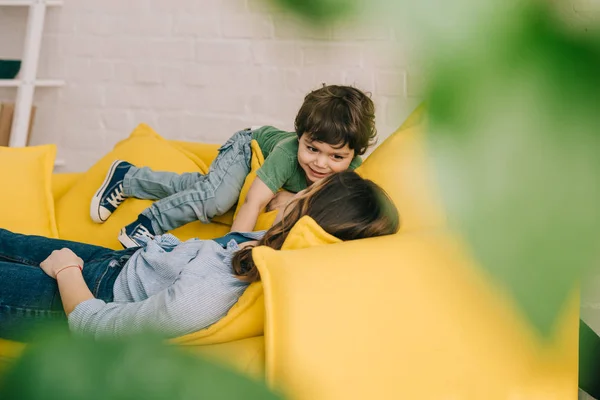 Petit garçon jouer avec la mère fatiguée alors qu'elle se trouve sur le canapé jaune dans le salon — Photo de stock