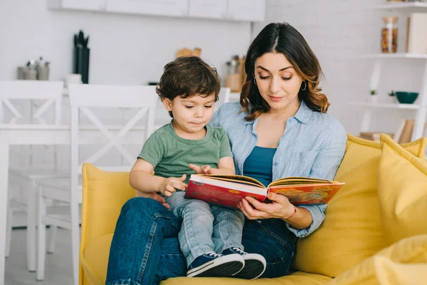 Mamá e hijo sentados en un sofá amarillo y leyendo un libro - foto de stock