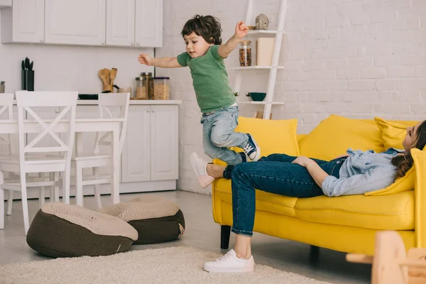 Kind amüsiert sich, während müde Mutter auf Sofa liegt — Stockfoto