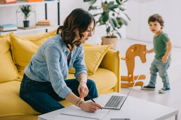 Kind schaut, wie Mutter mit Laptop im Wohnzimmer arbeitet — Stockfoto