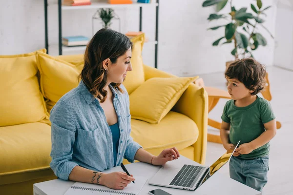 Gamin debout près de mère alors qu'elle travaille avec ordinateur portable — Photo de stock