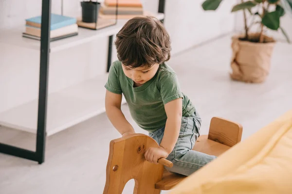Ребенок в зеленой футболке играет с деревянным качающимся конём — стоковое фото