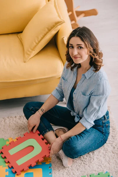 Overhead view of smiling woman in jeans sitting on carpet with puzzle mat — Stock Photo