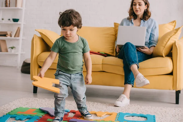 Lindo niño en camiseta verde jugando con la estera del rompecabezas, mientras que la madre usando el ordenador portátil - foto de stock