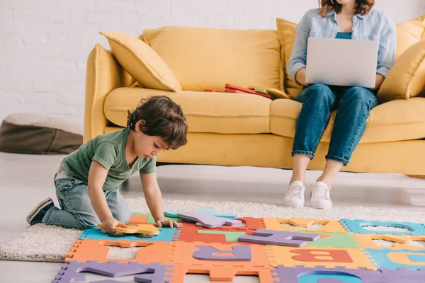 Cute child in green t-shirt playing with puzzle mat while mother using laptop — Stock Photo