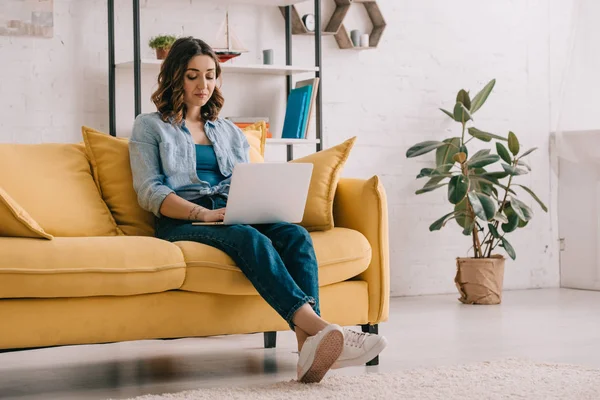 Attractive freelancer sitting on yellow sofa and using laptop — Stock Photo