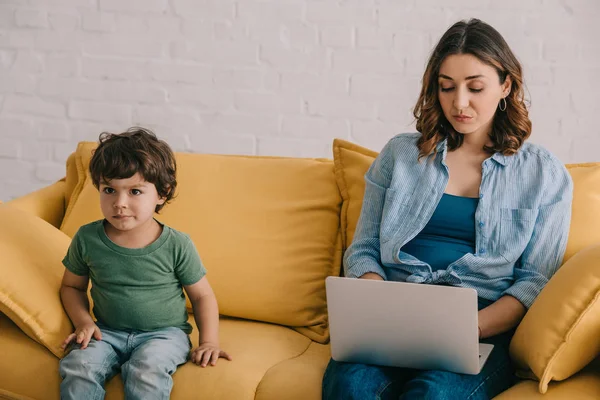 Mignon enfant assis sur le canapé tandis que la mère en utilisant un ordinateur portable — Photo de stock