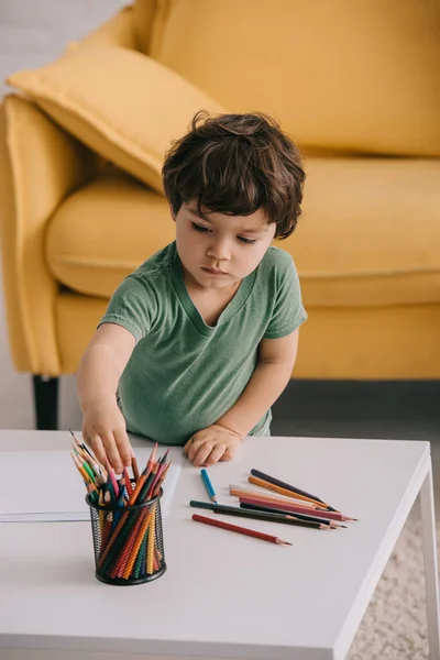 Ребенок в зеленой футболке с цветными карандашами и бумагами в гостиной — стоковое фото