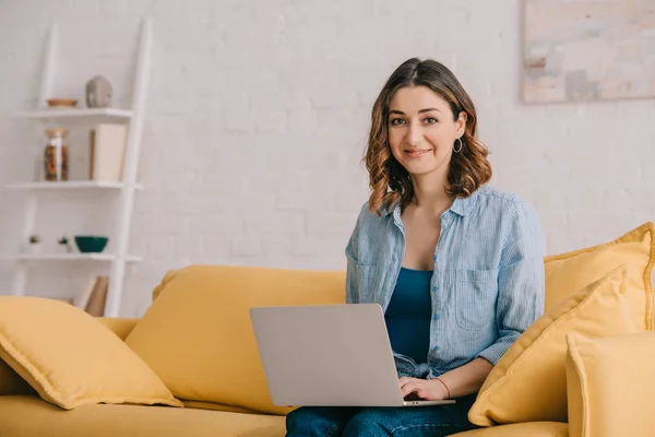 Attractive smiling freelancer sitting on yellow sofa and using laptop — Stock Photo