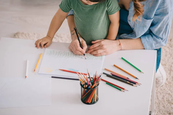 Vista recortada del dibujo de madre e hijo con lápices de color en la sala de estar - foto de stock