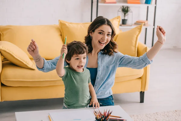 Animado mãe e filho com lápis de cor acenando as mãos na sala de estar — Fotografia de Stock