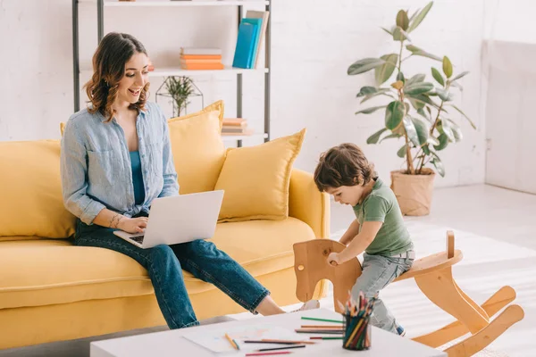 Kid sitting on rocking horse while mother working with laptop in living room — Stock Photo