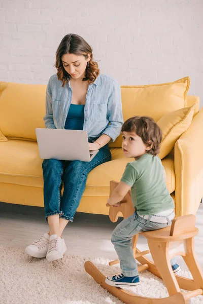 Kind sitzt auf Schaukelpferd, während Mutter im Wohnzimmer mit Laptop arbeitet — Stockfoto