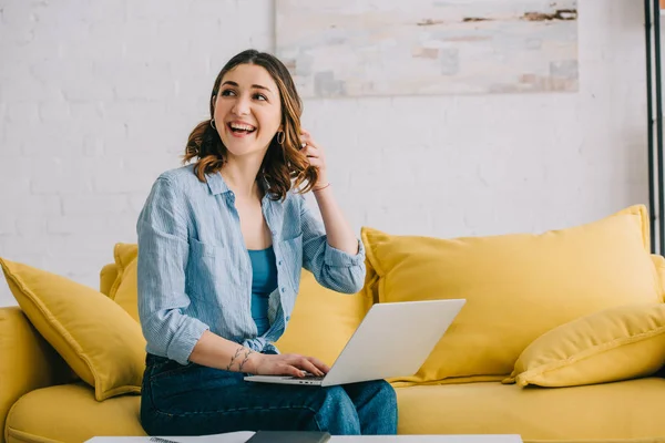 Smiling pretty woman sitting on yellow sofa with laptop — Stock Photo