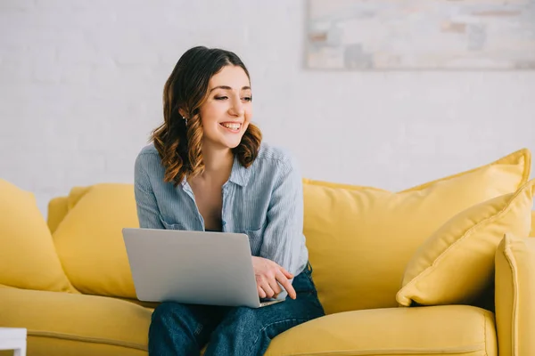 Smiling pretty freelancer sitting on yellow sofa with laptop — Stock Photo