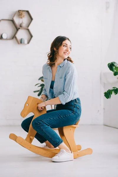Joyful woman in jeans sitting on rocking horse and looking away — Stock Photo