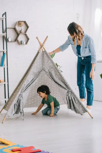 Visão de comprimento total da mulher em pé perto do filho em wigwam cinza — Fotografia de Stock