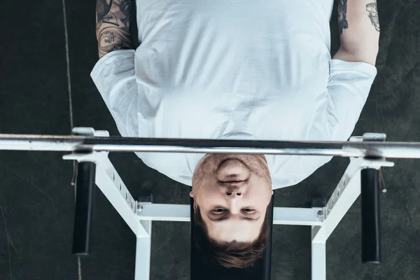 Вид зверху татуйований чоловік з надмірною вагою дивиться на камеру під час тренувань з барбелом у спортзалі — стокове фото